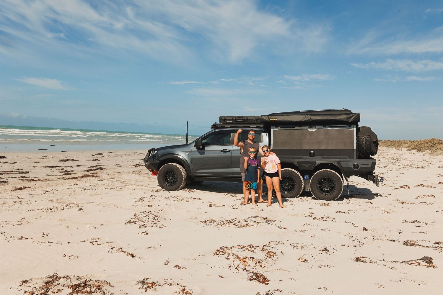 Black off-road vehicle with roof tent parked on sandy beach with ocean and blue sky in background.