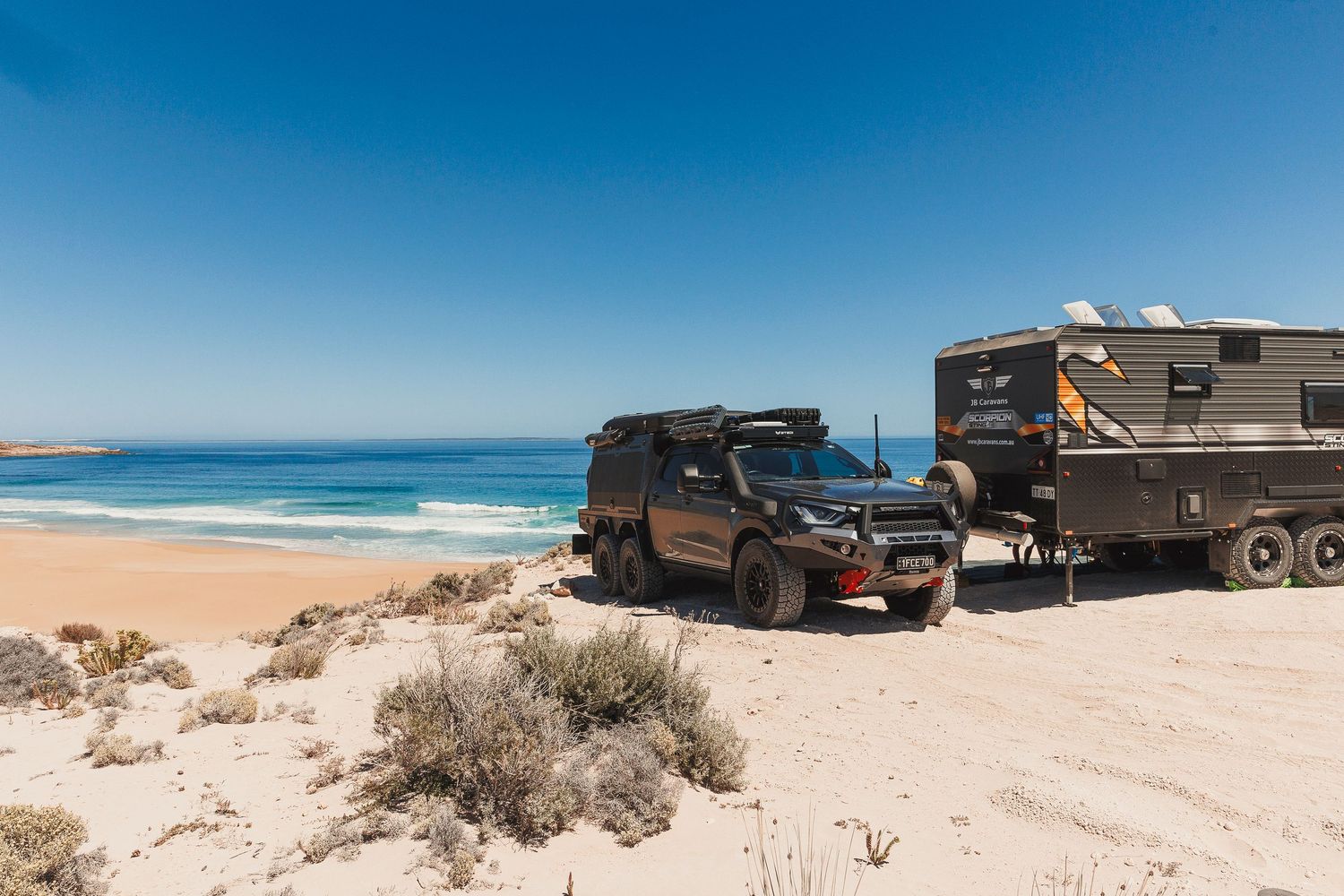 A panoramic view of large military trucks and vehicles parked along a pristine sandy beach with blue ocean waters.