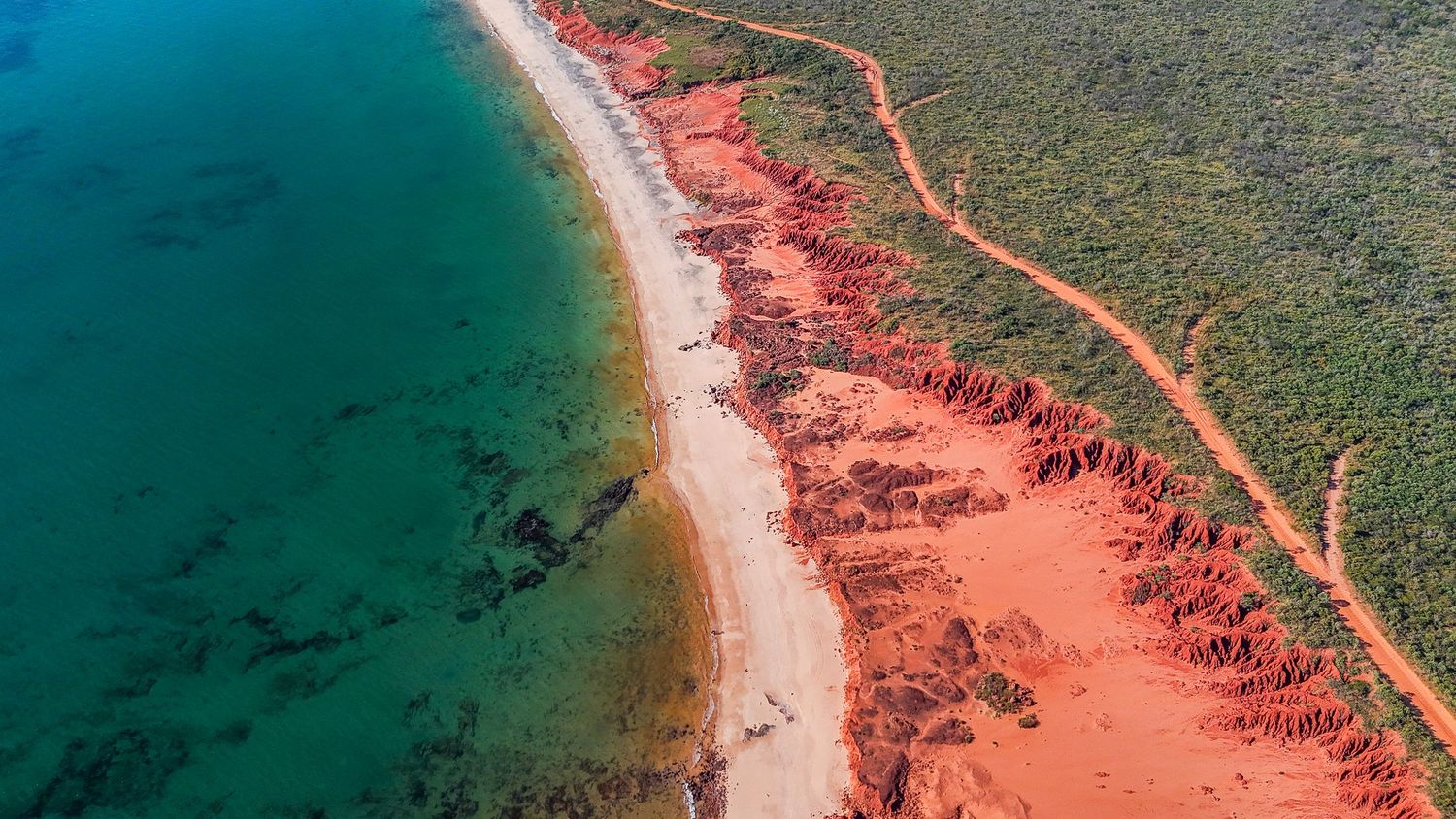 Aerial view of a winding coastal road along turquoise waters and coral-colored cliffs in a tropical paradise.