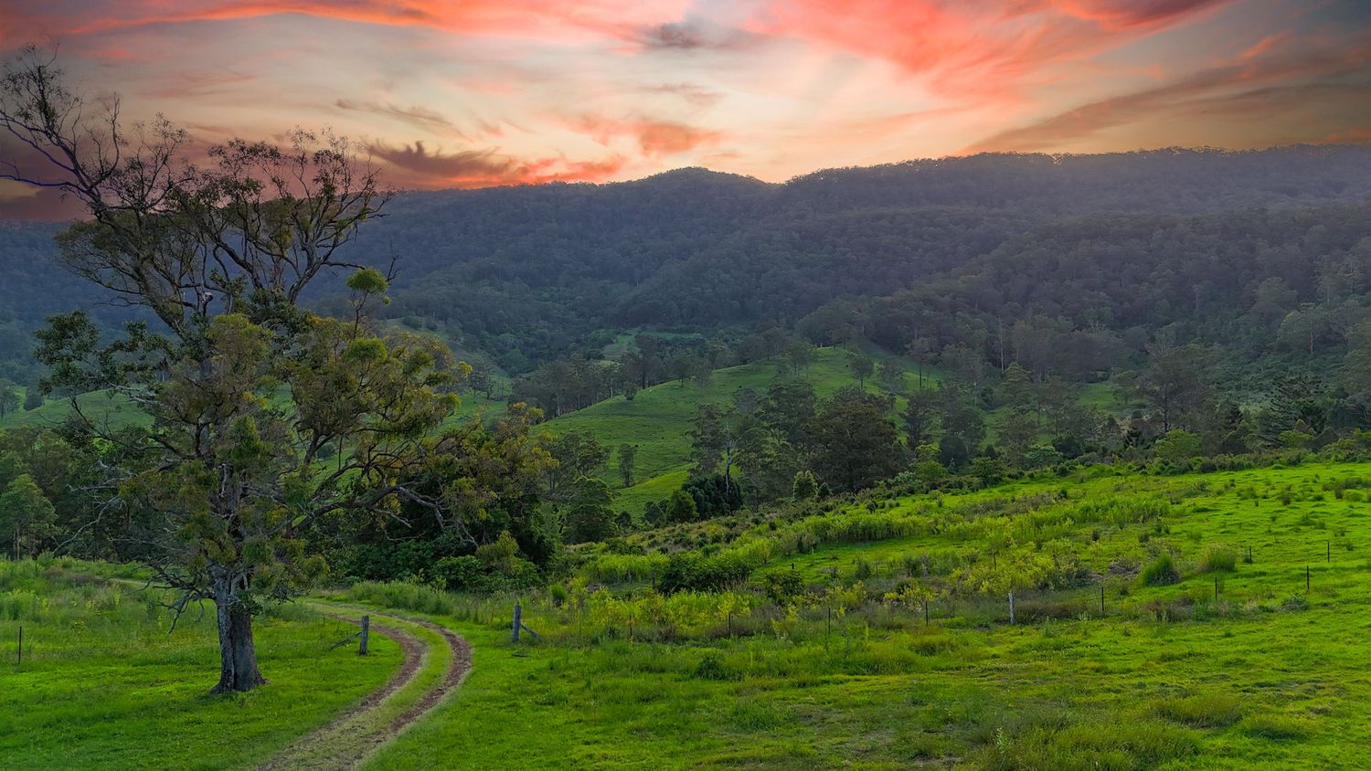 Dramatic sunset sky over rolling green hills and mountains with a winding path.