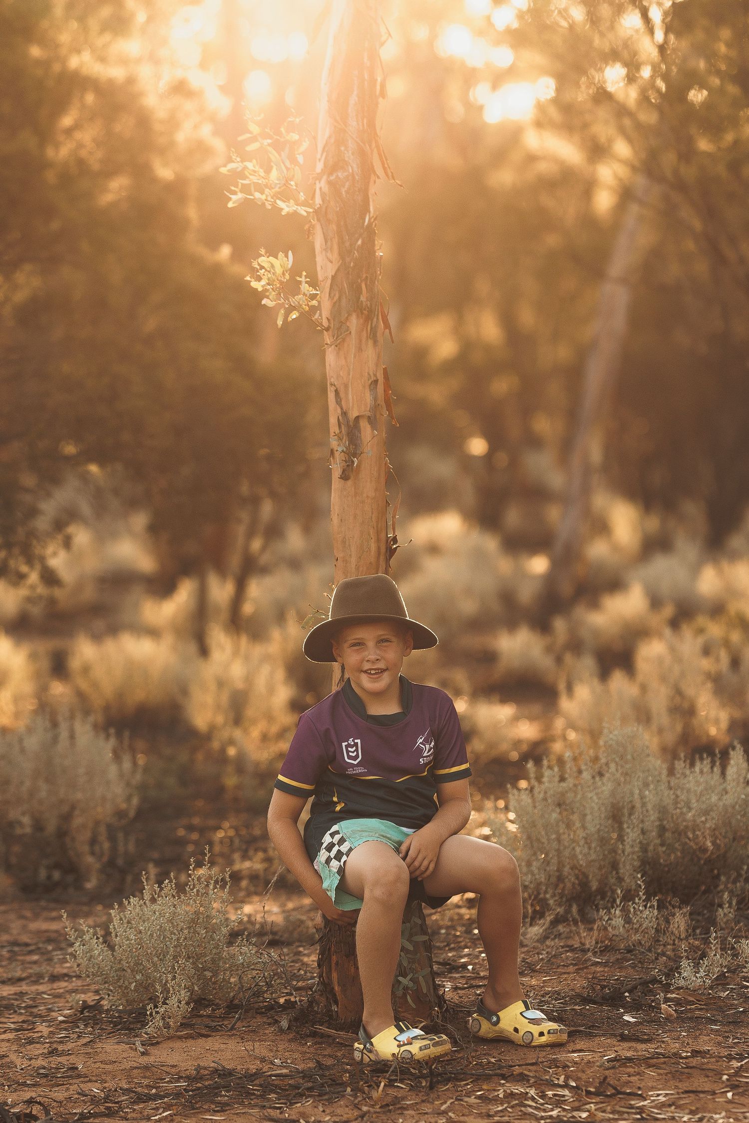 Young child in a brown hat sits and smiles among sagebrush at sunset in a natural outdoor setting.
