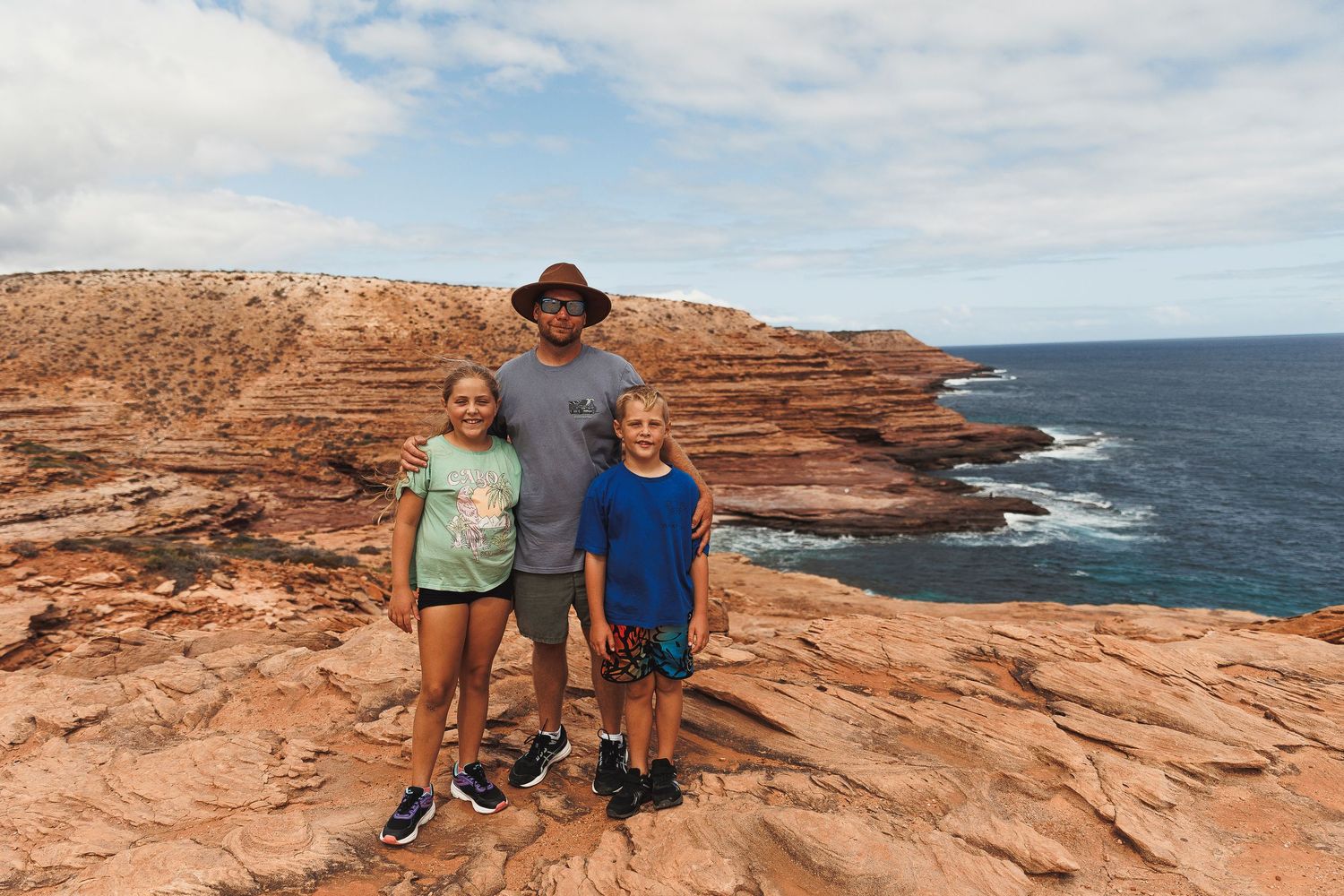 Hikers stand on red sandstone cliffs overlooking the rugged ocean coastline on a sunny day.