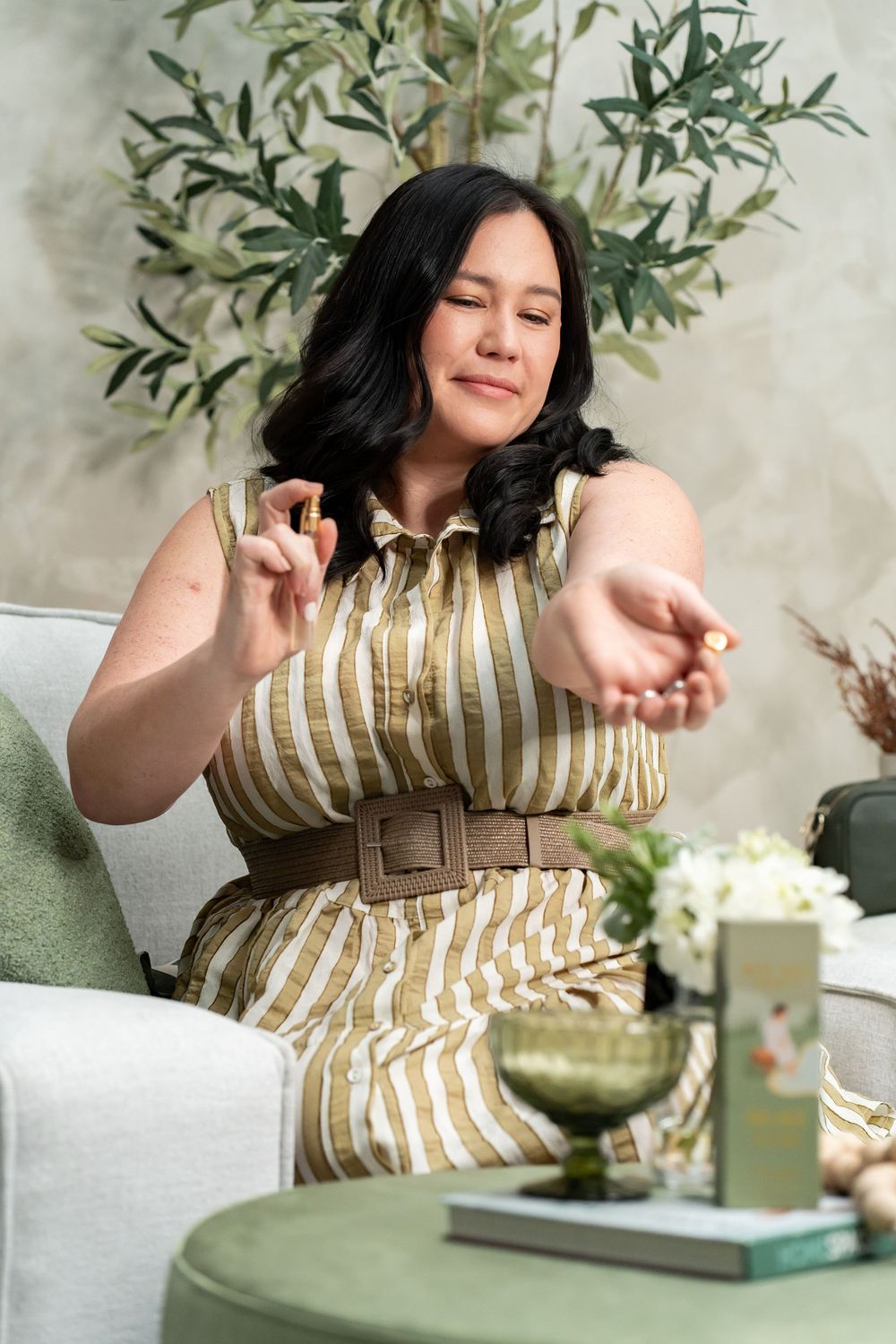 A person in a striped dress with a brown belt sits on a couch with olive plants in the background.