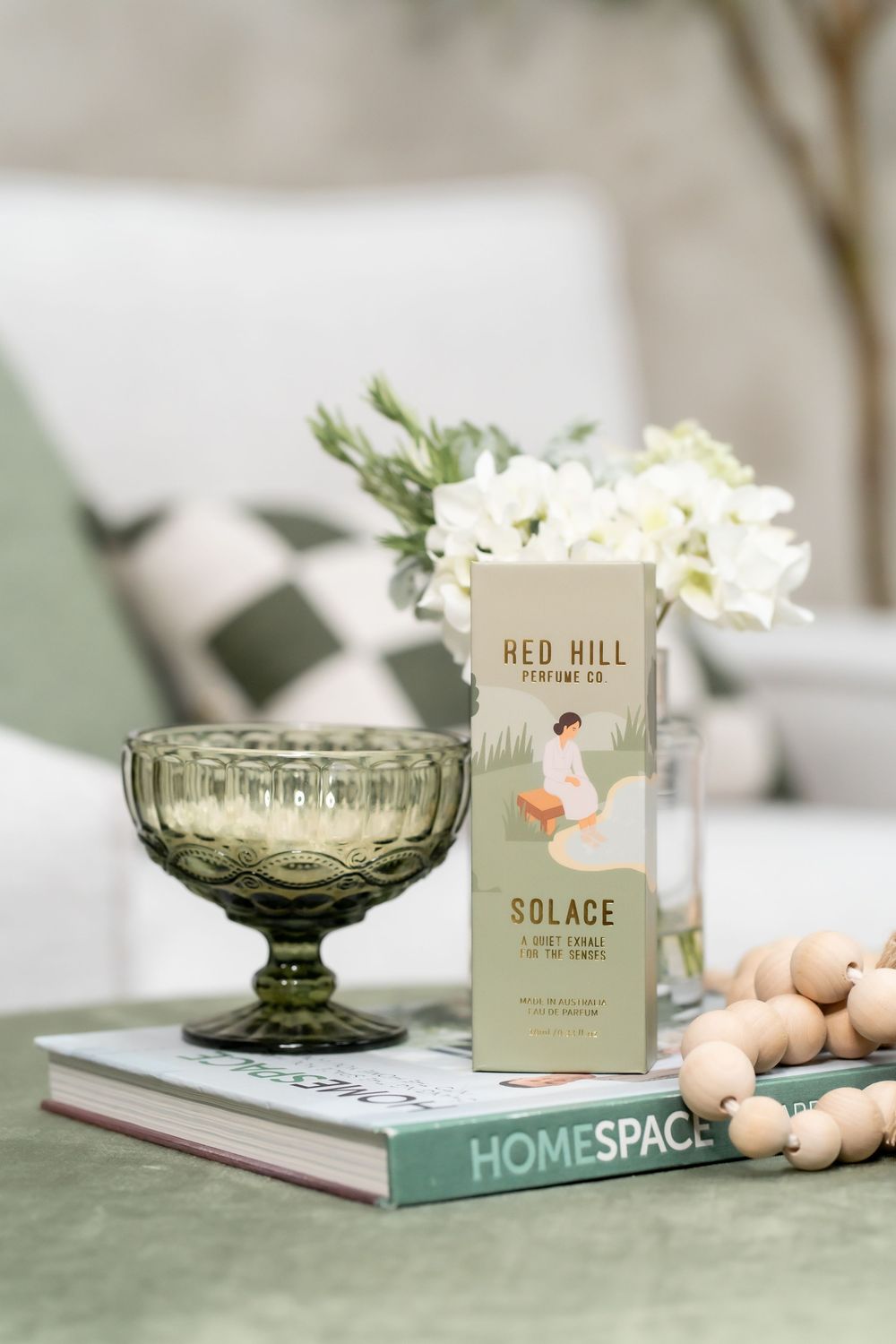A green vintage glass compote dish with white flowers sits beside candles and books in a soft, minimalist styling setup.