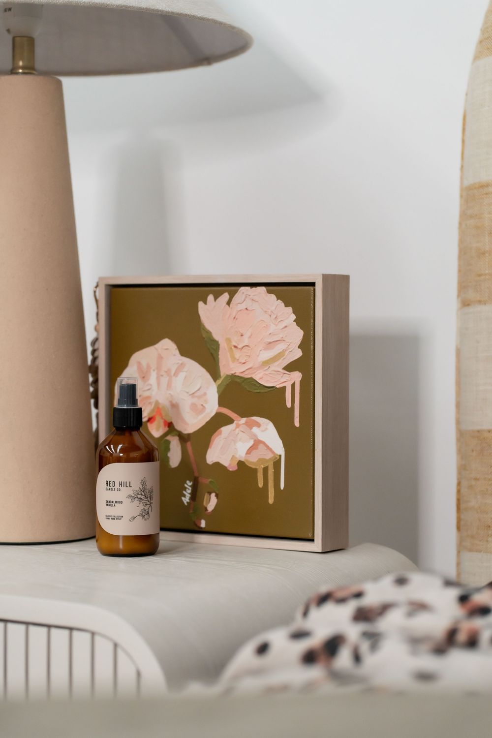 Delicate pink floral art print with an amber bottle displayed against a neutral wall under a table lamp.