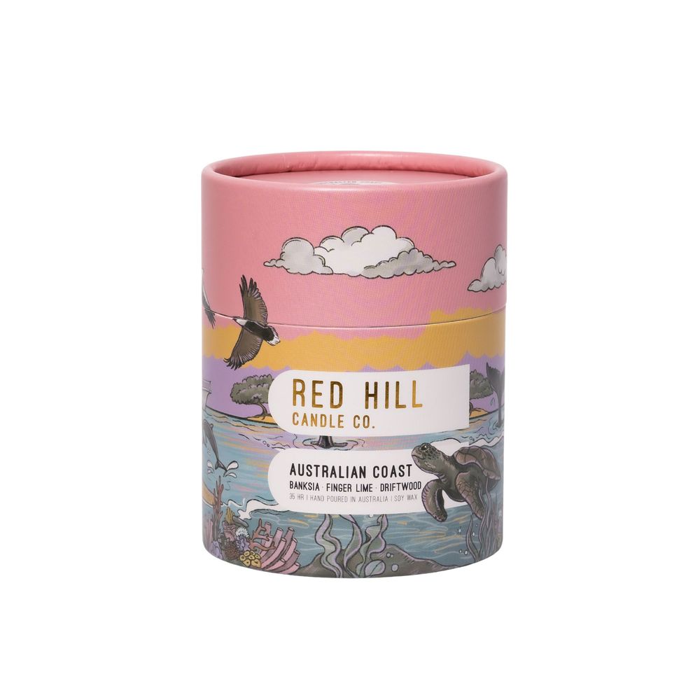 Pink candle in cylindrical glass jar with decorative coastal landscape scene printed on exterior showing clouds and waves.