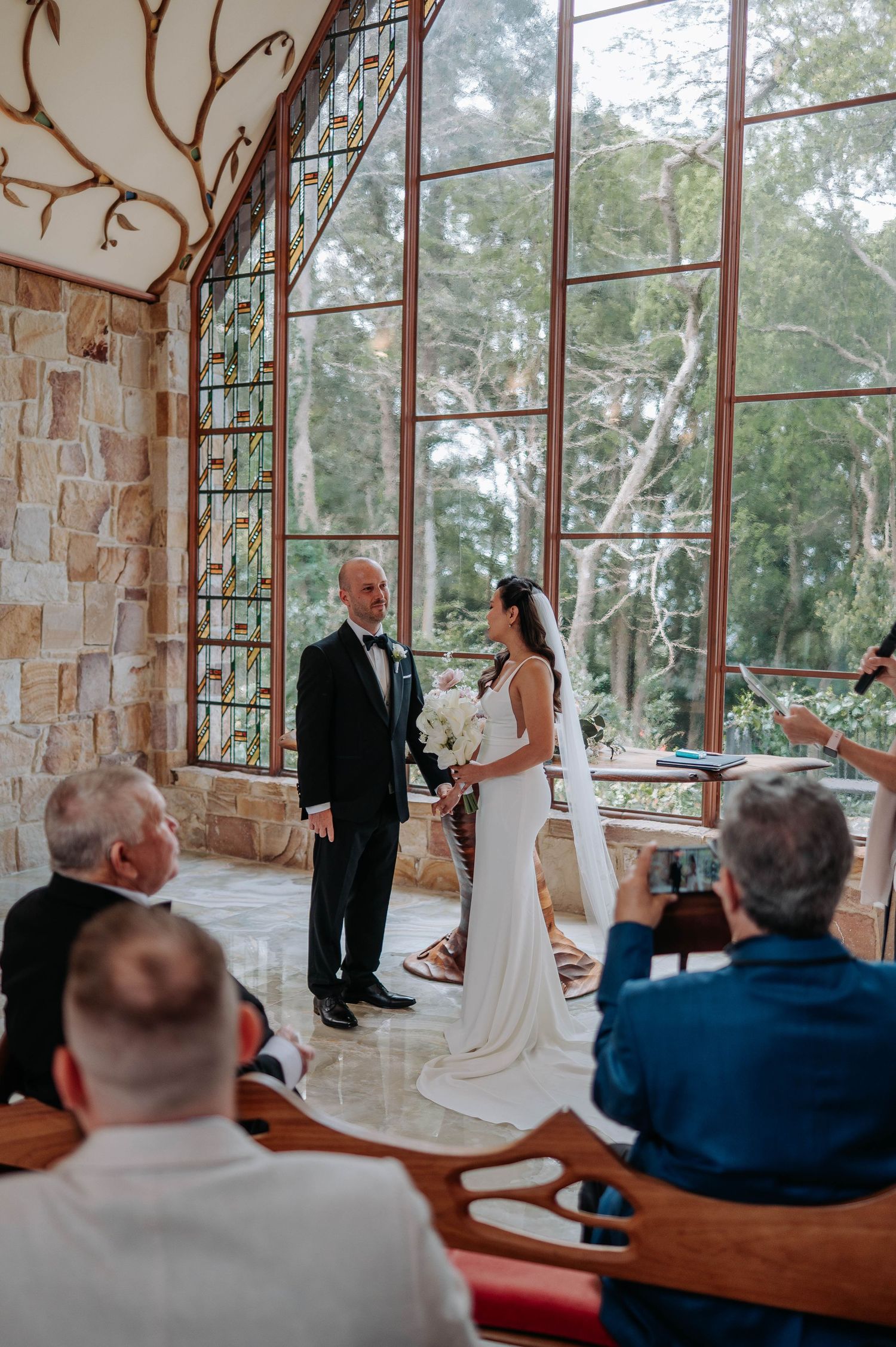 An elegant indoor wedding ceremony takes place in a chapel with floor-to-ceiling windows overlooking lush greenery.