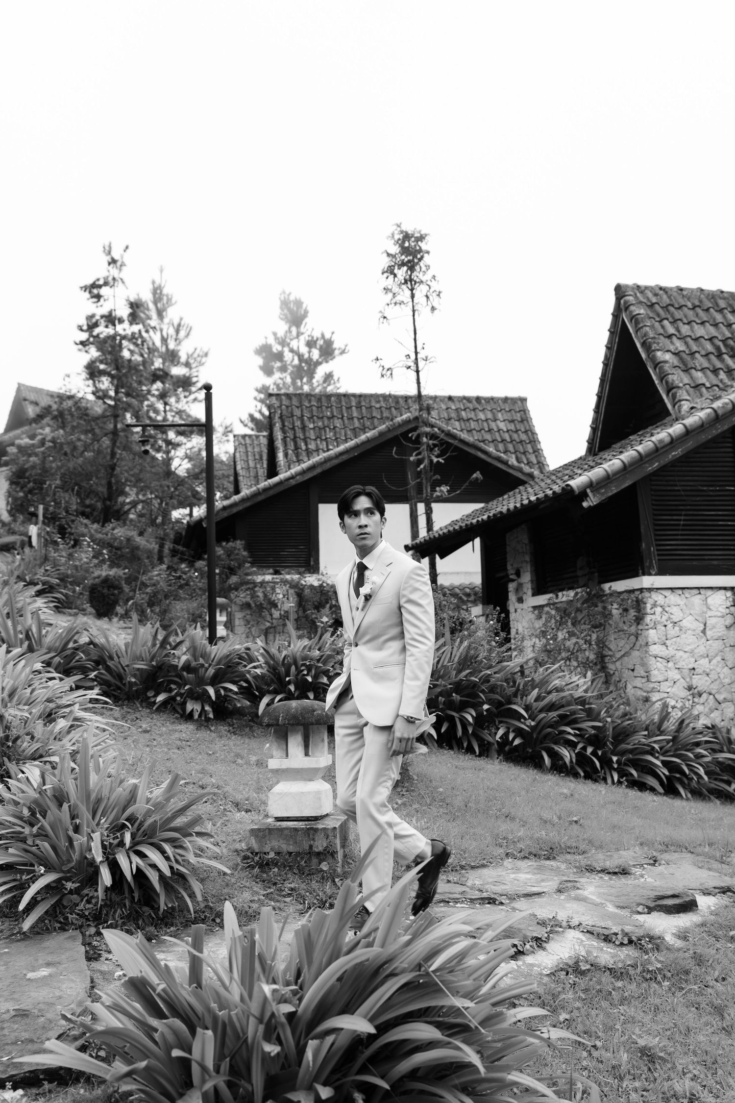 Black and white photo of groom in white suit posing in lush garden with stone cottages in background.