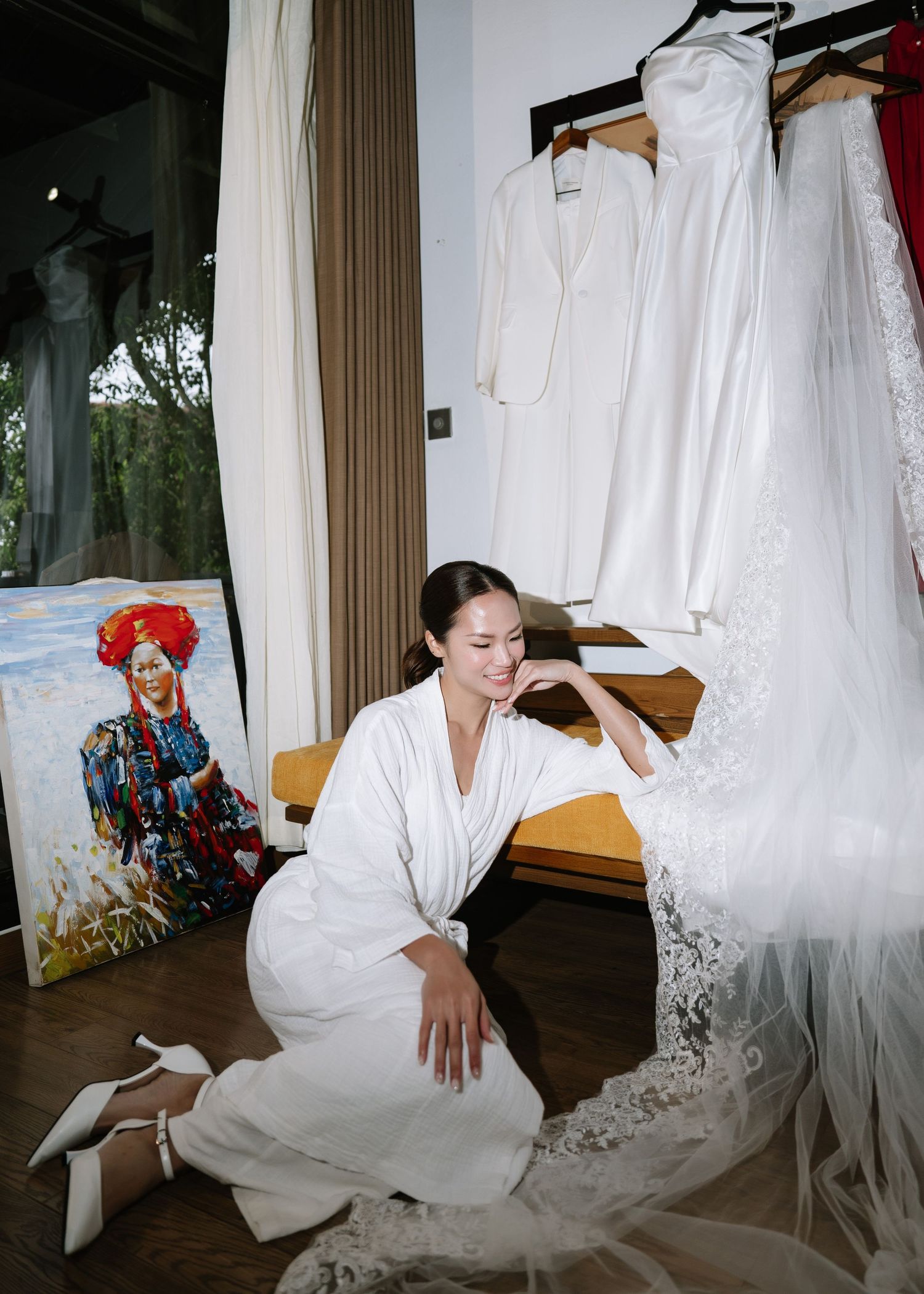 Bride in white robe kneeling beside lace wedding dress with colorful portrait painting nearby.