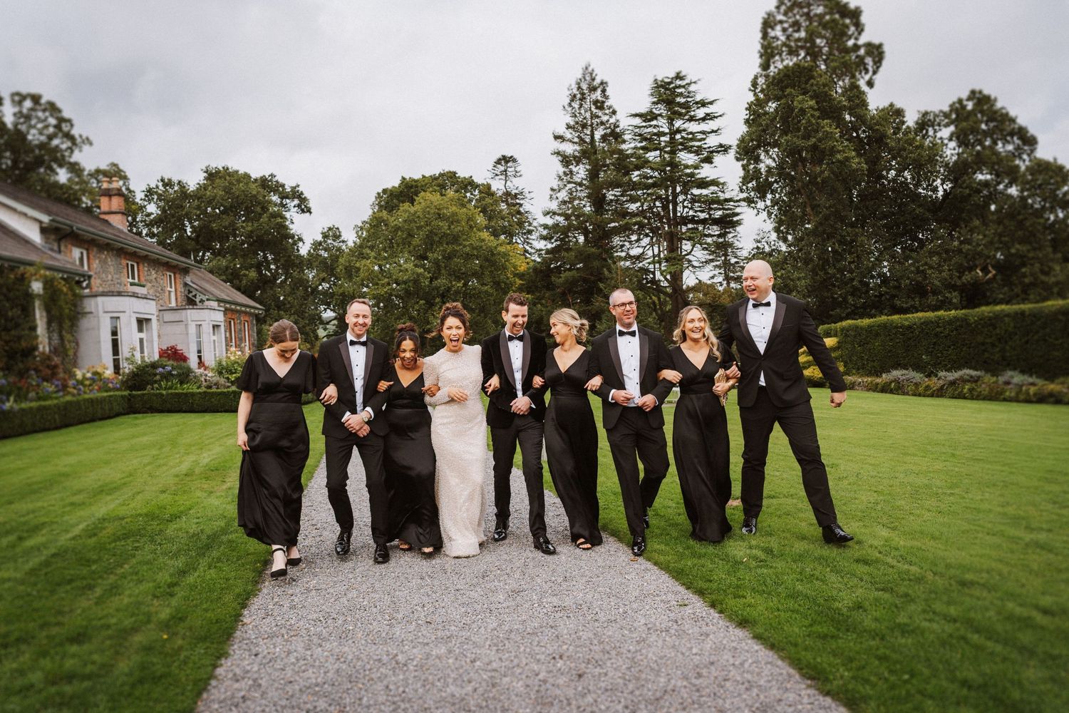 Wedding party members pose in formal attire on a lush green lawn in front of a classic estate building.