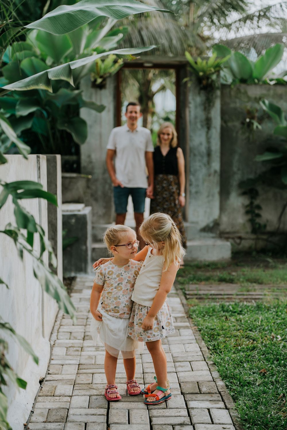 Two young children share a sweet moment on a tropical garden path with lush greenery.