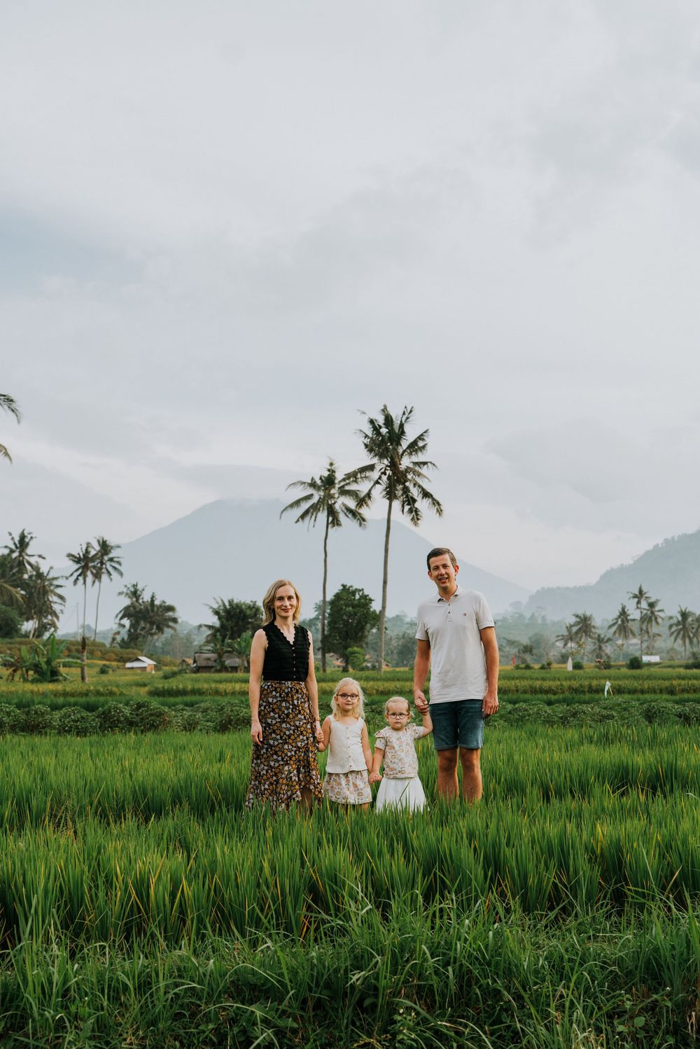 Family standing in a vibrant green rice field with palm trees and mountains in the distance.