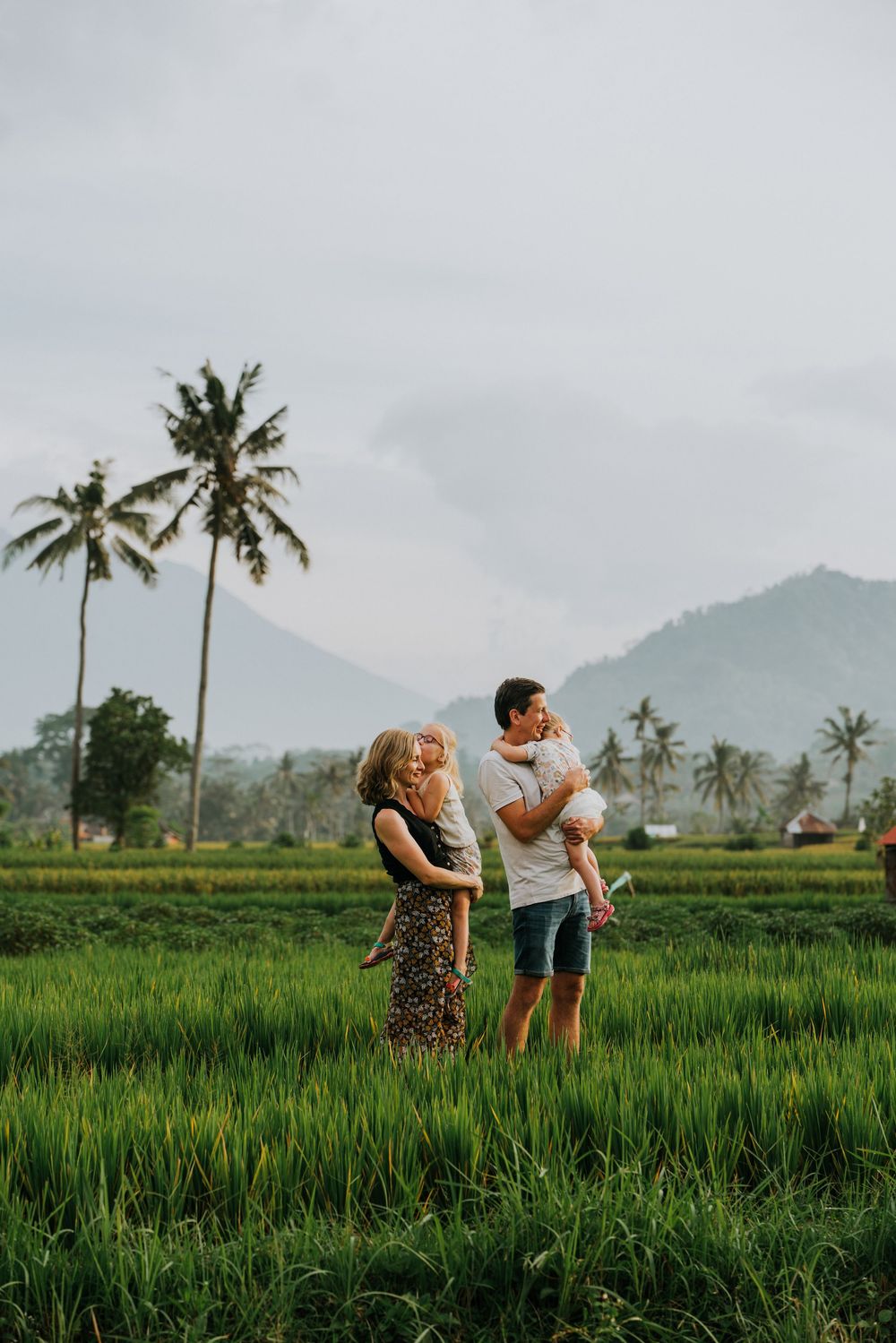 Family enjoying sunset in lush rice fields with palm trees and mountains in the distance.