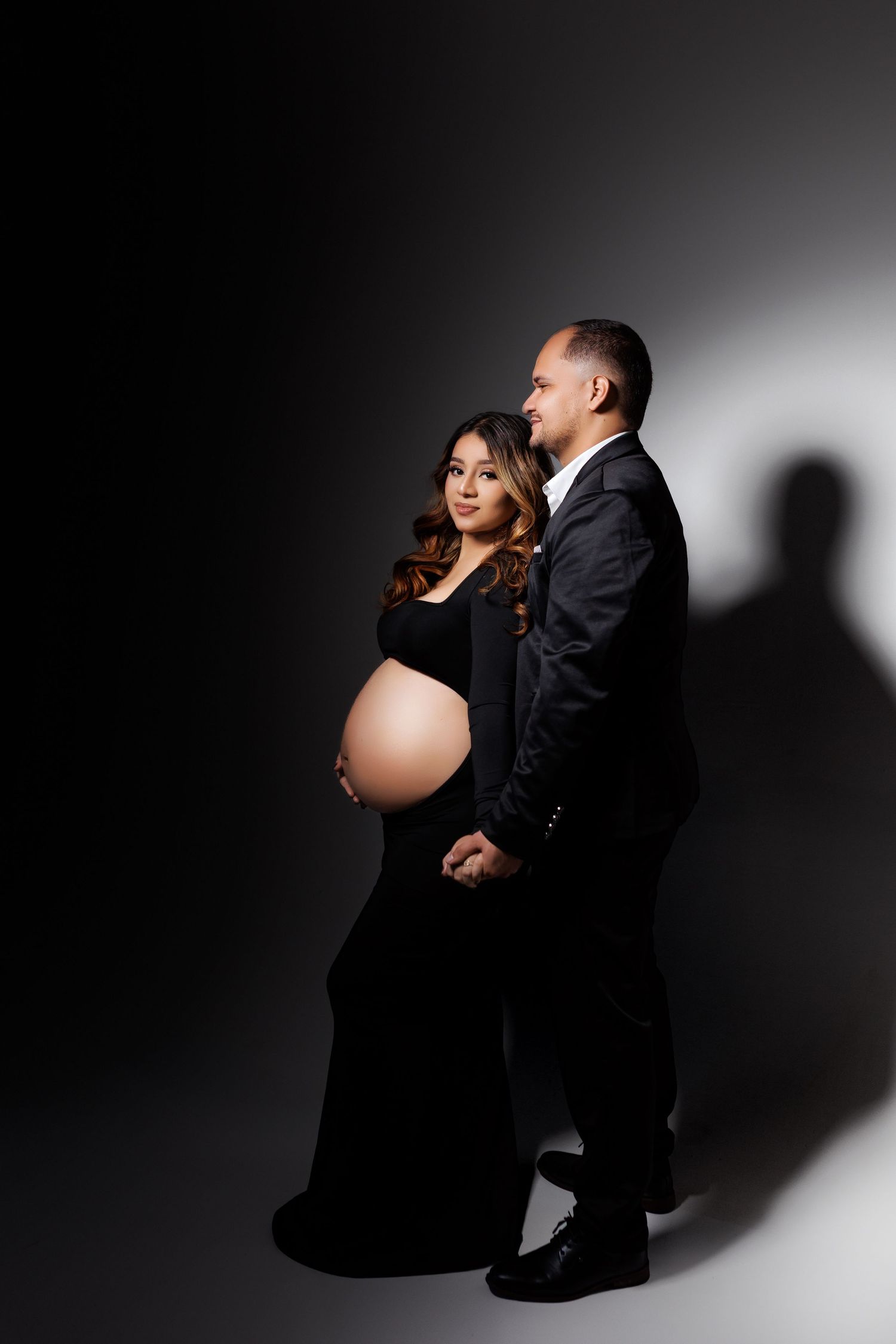 Artistic maternity photo featuring couple in formal wear against dark backdrop with side lighting.
