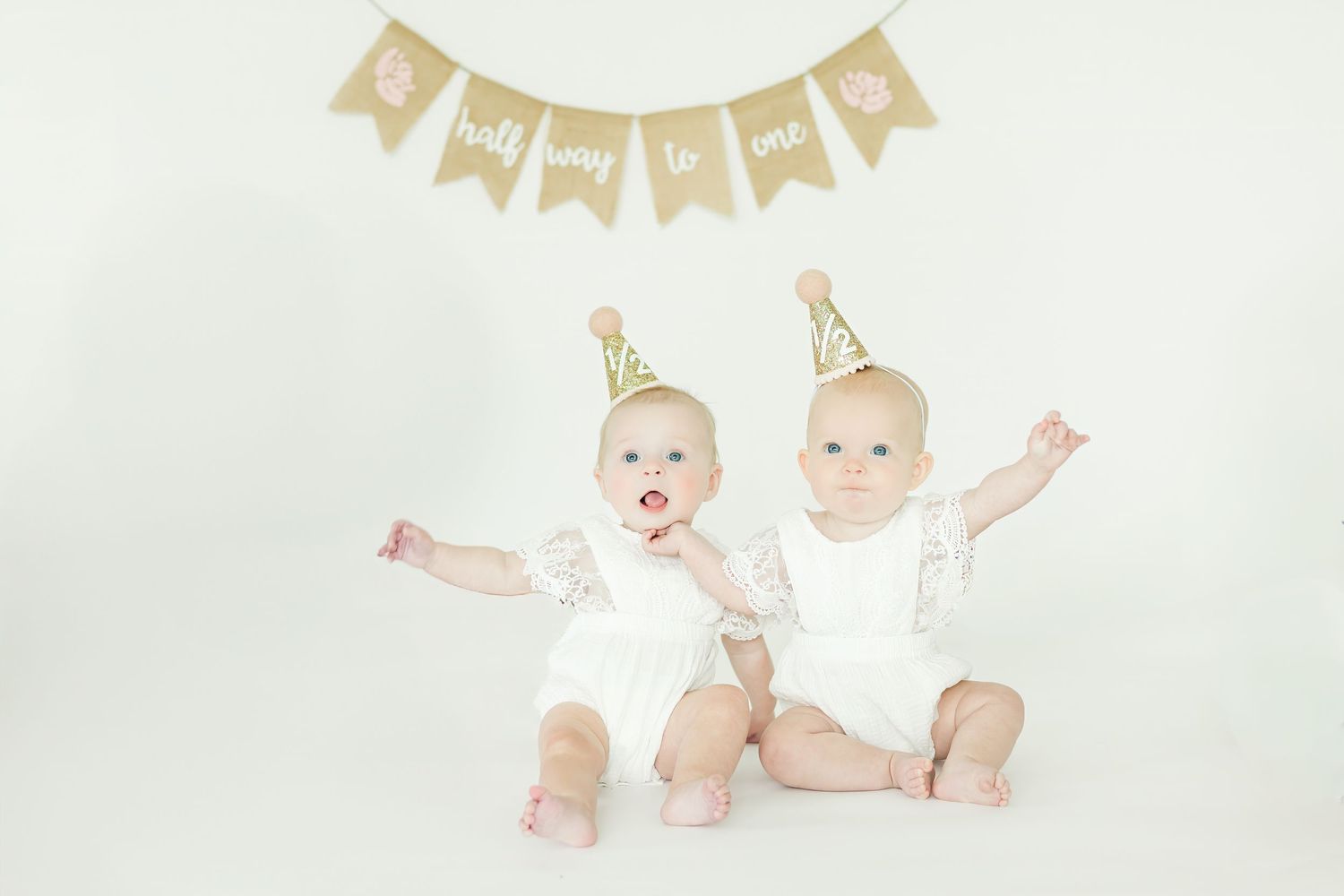 Two infants wearing white outfits and party hats sit together under a decorative banner.
