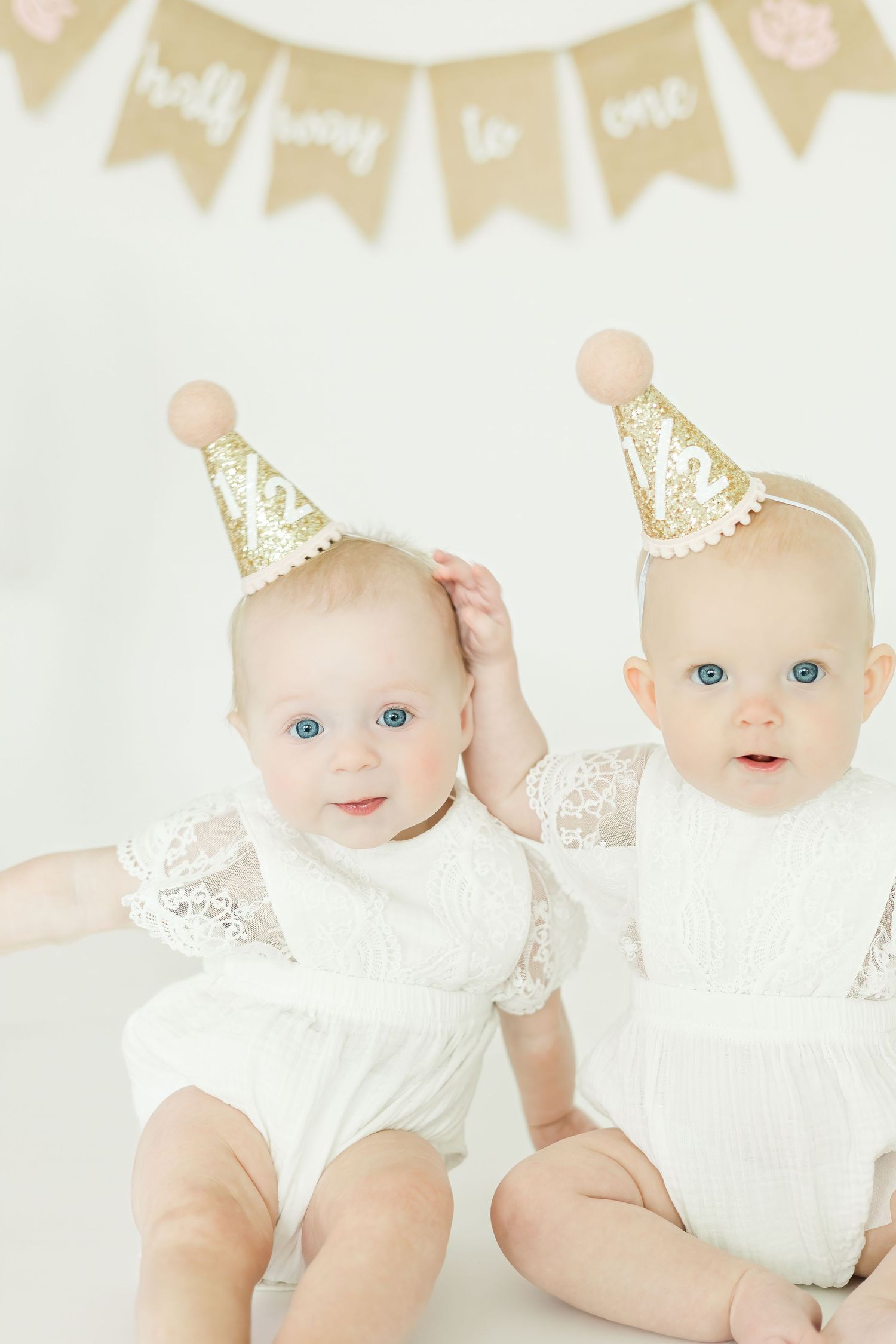 Two babies in white lace outfits and gold party hats pose for a celebratory photo.