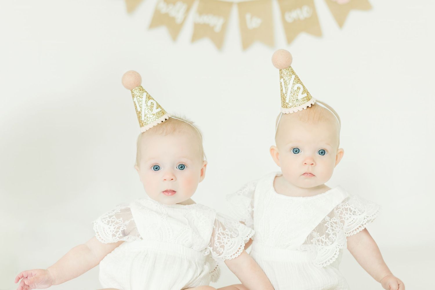 Two infants in matching white lace outfits and gold party hats sit together under a banner.