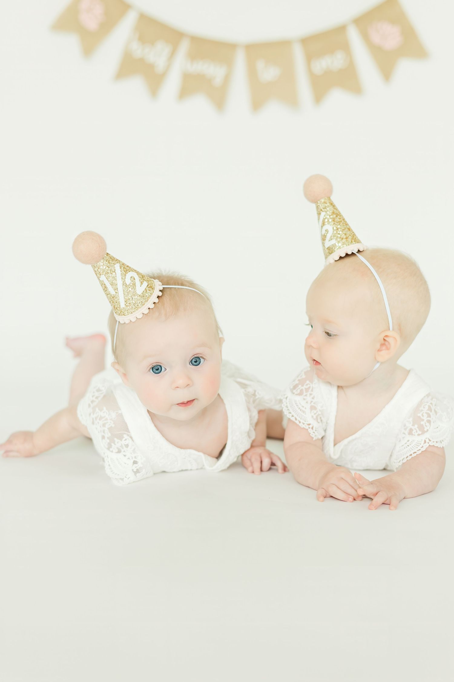 Two babies in white lace rompers and gold party hats lay on a light background.