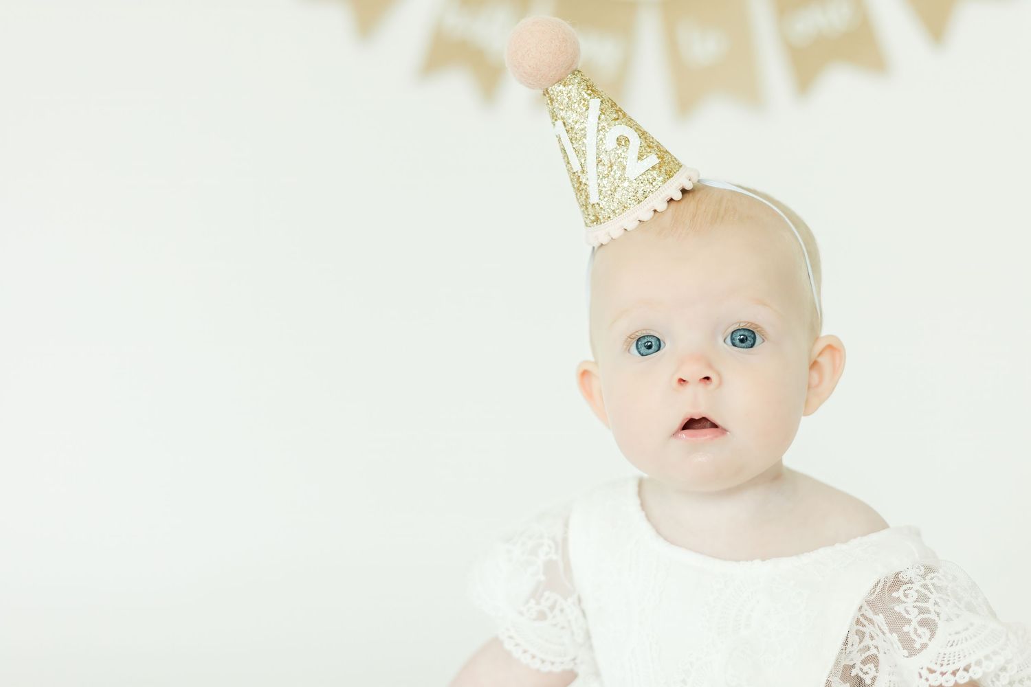 Baby wearing white lace outfit and sparkly gold party hat poses against light background.