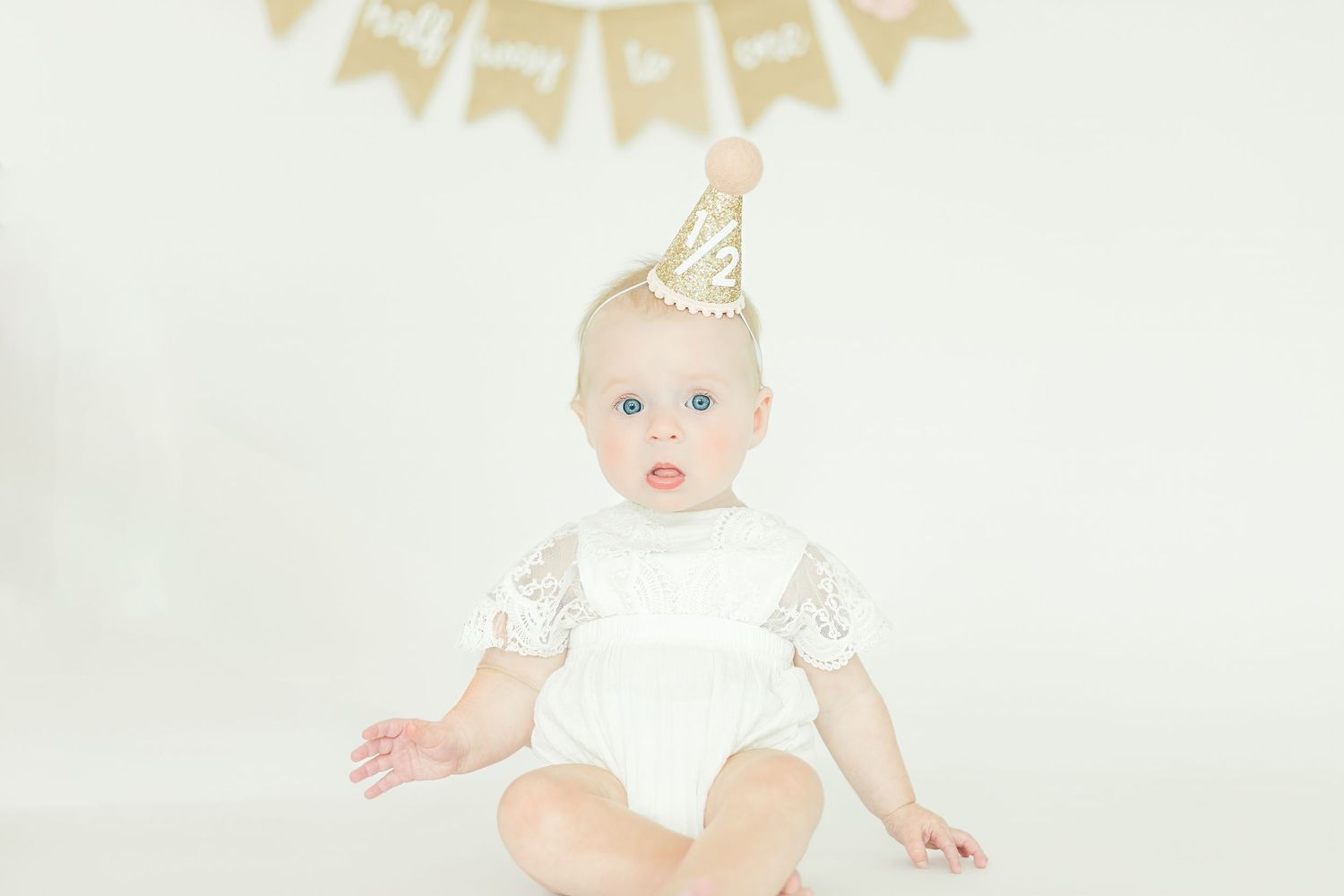 Baby in white lace outfit and gold party hat sits under decorative banner.