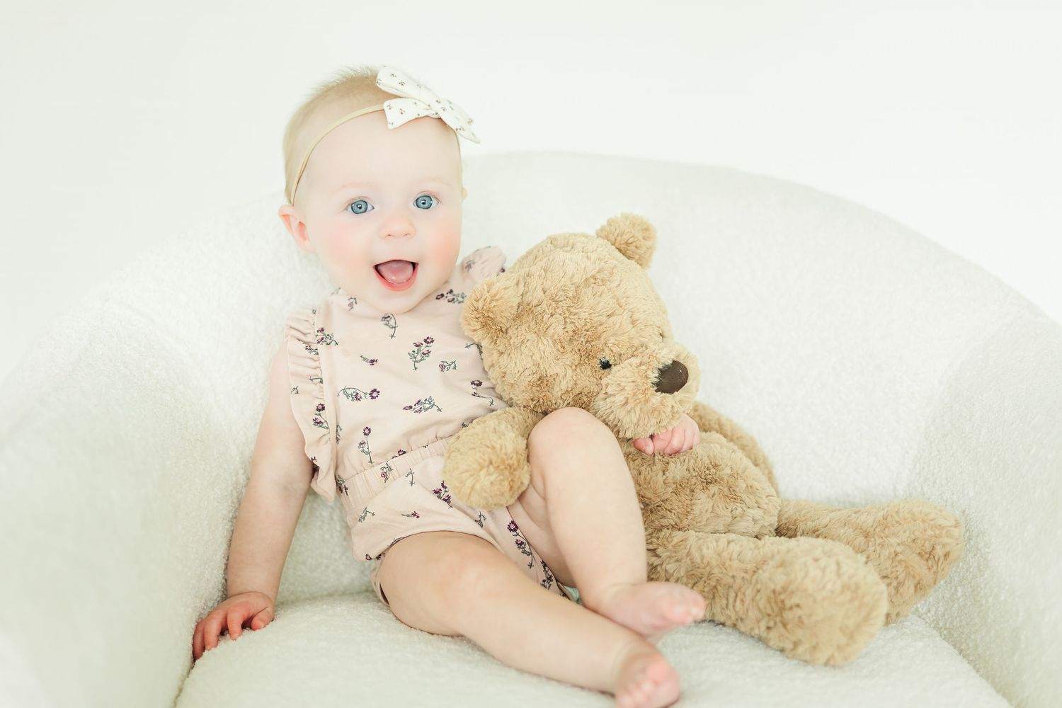 Smiling baby in floral outfit sits with teddy bear on white couch.