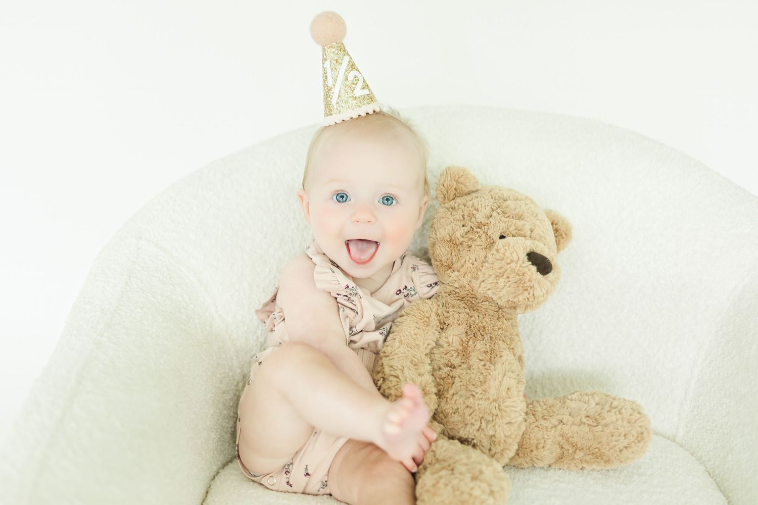 Happy baby wearing party hat poses with brown teddy bear on white furniture.