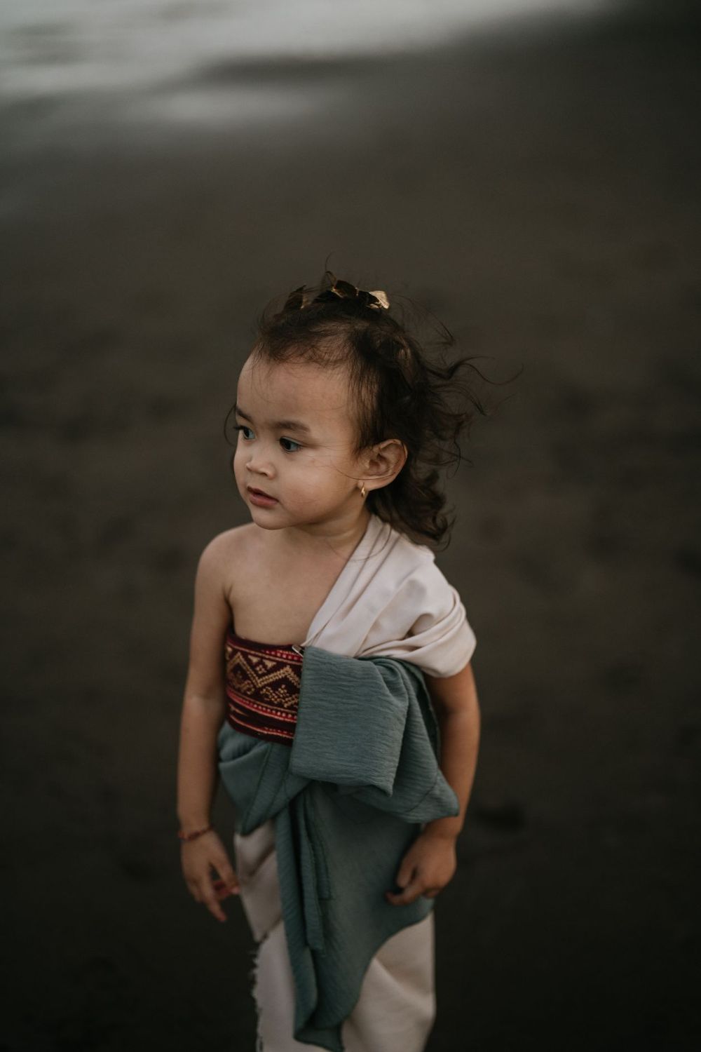 Young child in white dress with teal fabric on a dark beach at dusk.