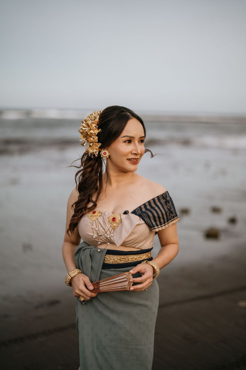 Woman in sage dress with gold hair accessories poses on moody beach at sunset.