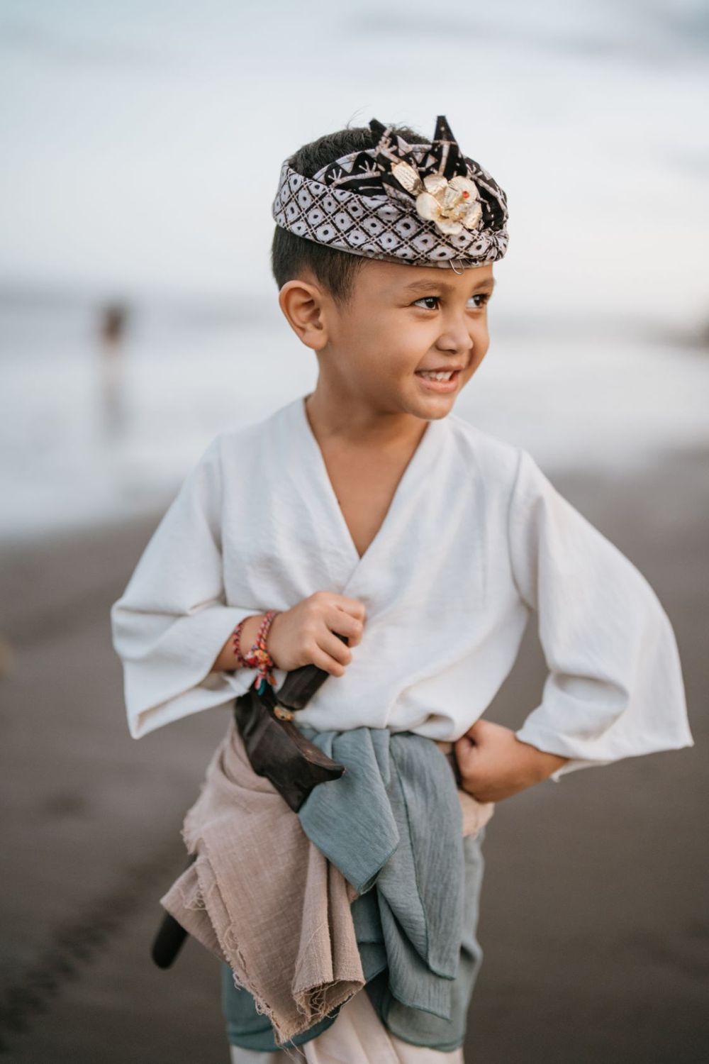 Child wearing traditional white clothing and headband smiles while playing on beach.