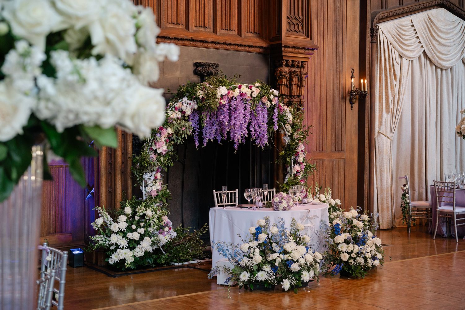 Elegant wood-paneled ballroom decorated with white floral arrangements and drapery for a luxurious wedding reception.
