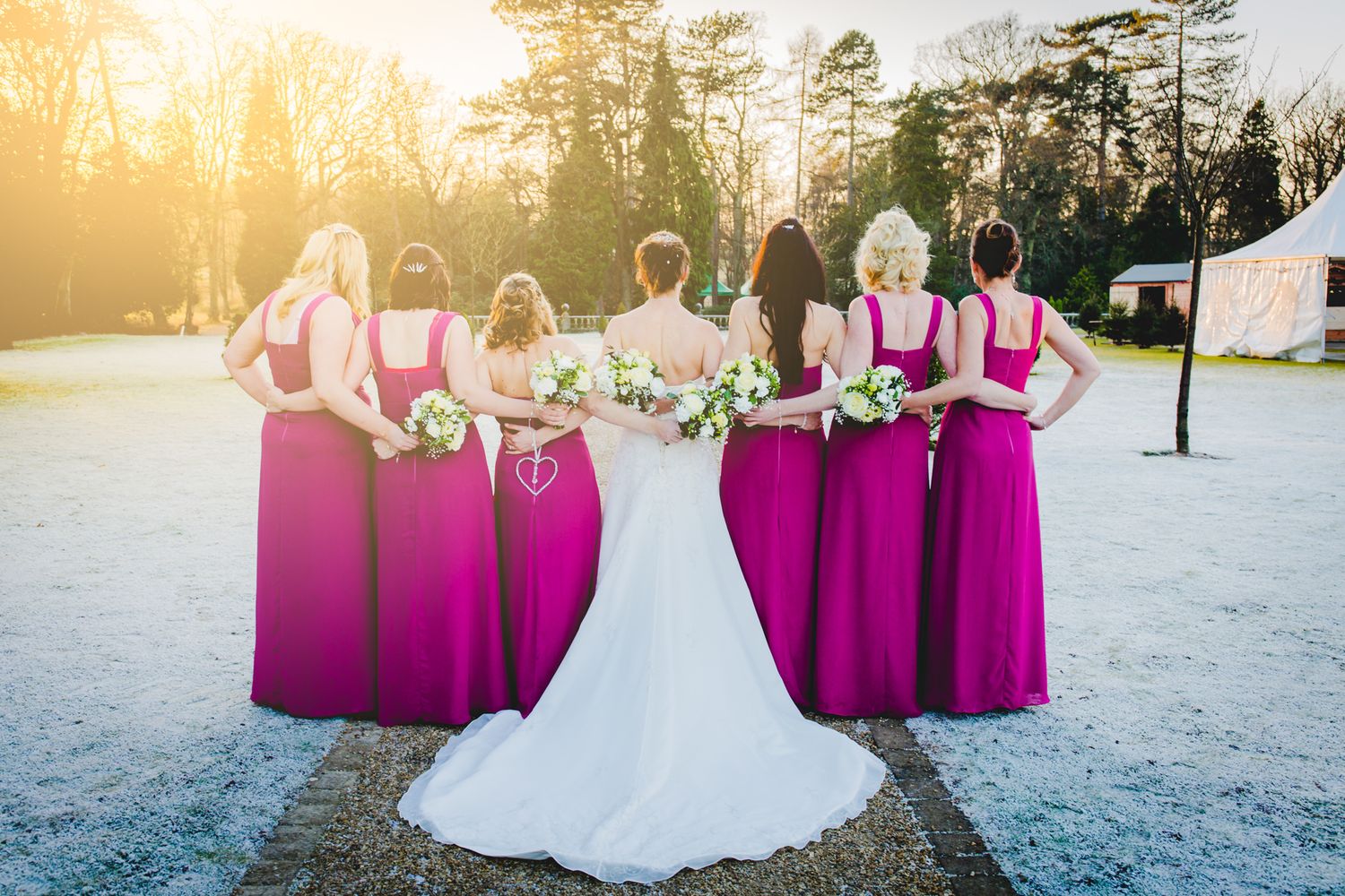 Bridesmaids in magenta dresses stand with bride in white gown on snowy ground during golden sunset.