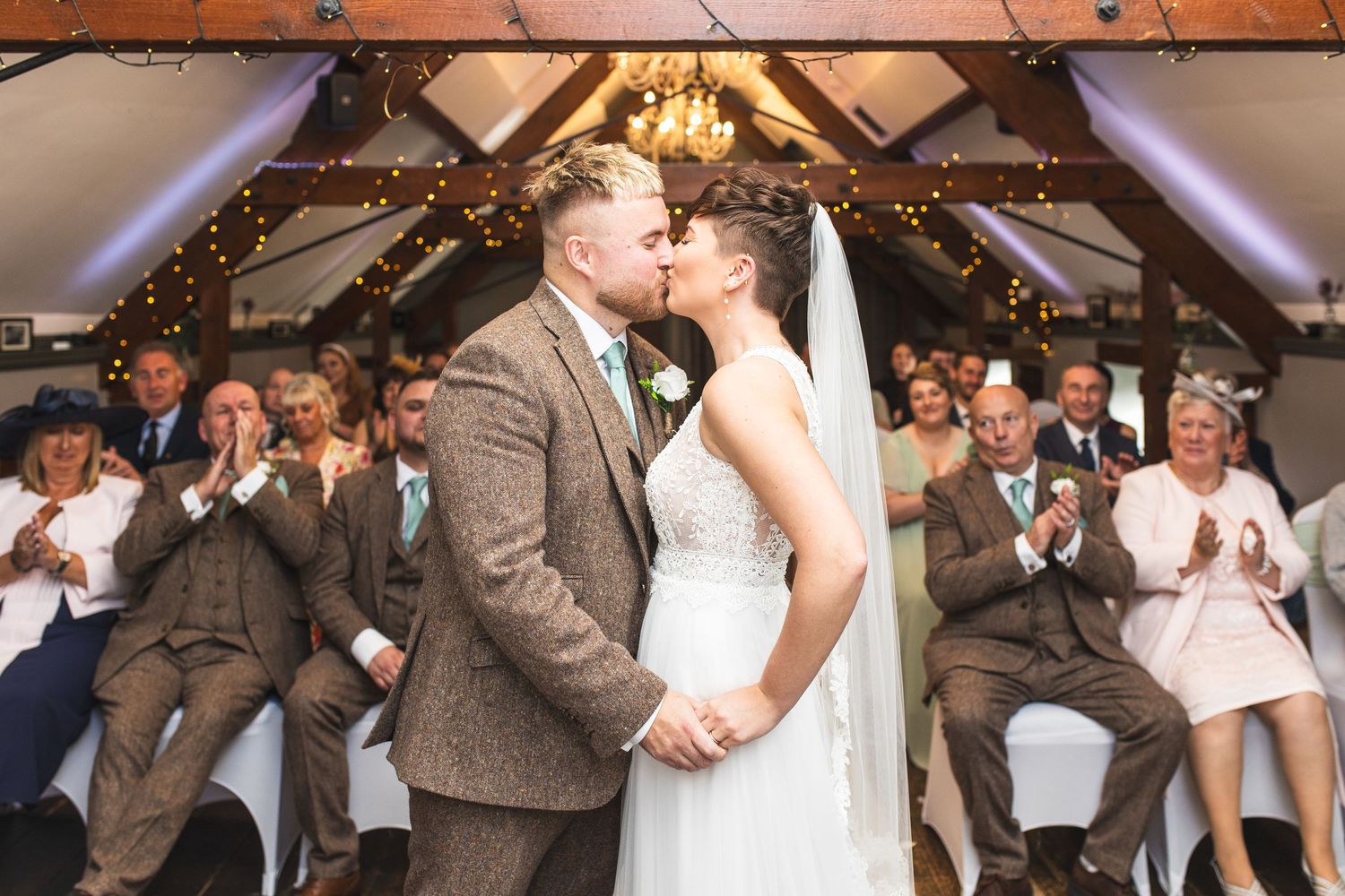 Wedding party celebrates outside a rustic venue with guests in brown suits and pastel dresses.