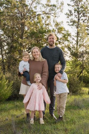 Family portrait in casual attire standing together on a grassy hillside with trees in background.