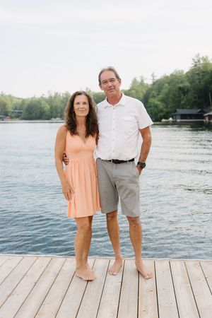 A couple stands together on a wooden dock overlooking a peaceful lake on a sunny day.