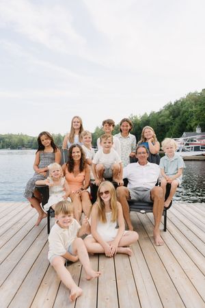 A large group sits casually arranged on a wooden dock by the water on a bright summer day.