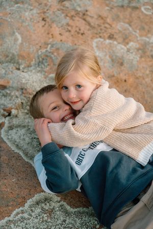 Two children in cozy sweaters cuddling on textured rock surface.