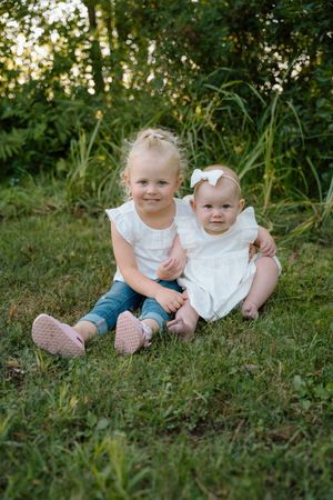 Two young children sitting together on grass wearing white and denim outfits.