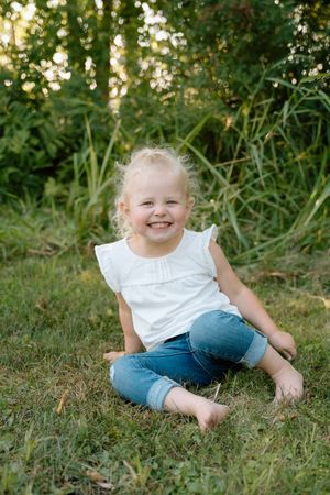 Child sitting on grass wearing white top and jeans during outdoor summer photoshoot.