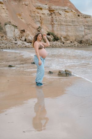 Beach maternity photoshoot with light blue dress against rocky cliffs and ocean waves.