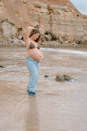 Expectant mother in blue dress walking along beach shoreline with rocky cliffs in background.