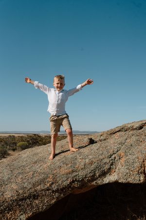 Child in white shirt and shorts balancing on rocky surface against blue sky.