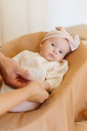 A baby in white clothing rests on warm-toned bedding while hands reach out tenderly.