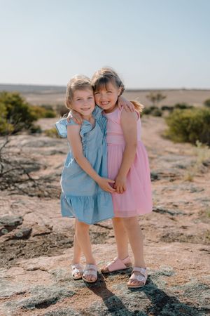 Two children in pink and blue dresses hugging on rocky terrain.
