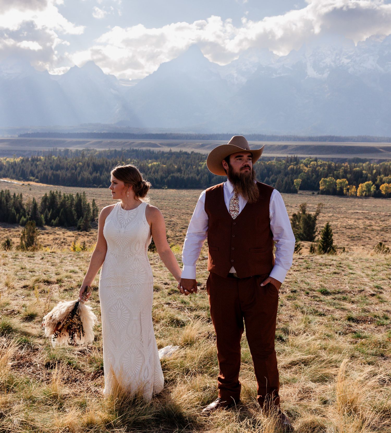 Couple in western wedding attire stands in a golden meadow with dramatic mountain views and cloudy skies at sunset. Jackson, Wyoming