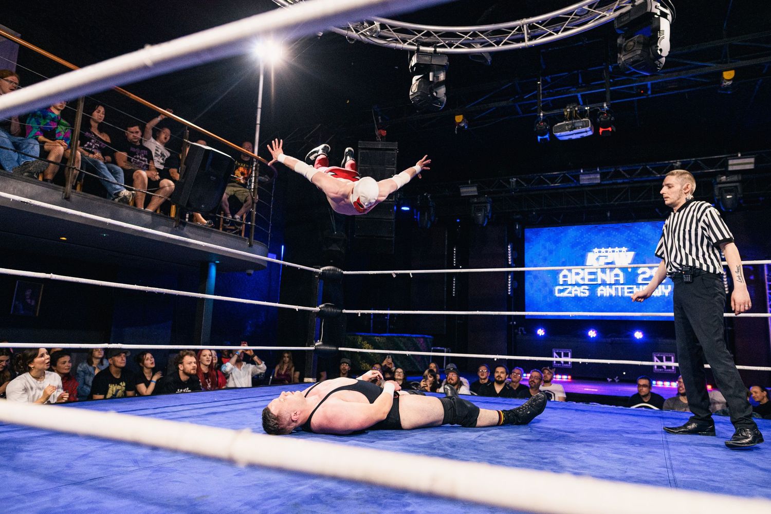 Wrestlers engage in acrobatic moves during an intense match in a brightly lit wrestling arena.