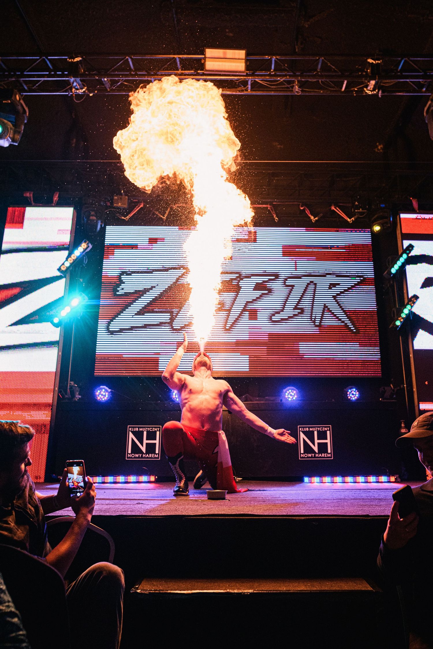 Professional wrestler breathes fire during dramatic entrance performance on wrestling stage with illuminated backdrop.