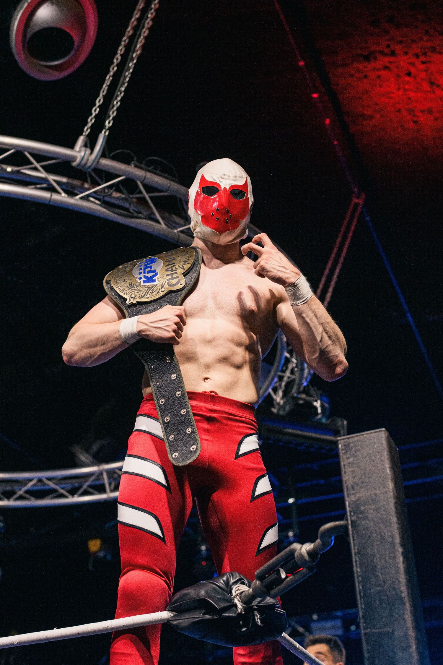 Masked wrestler in red attire displays championship belt in wrestling ring.