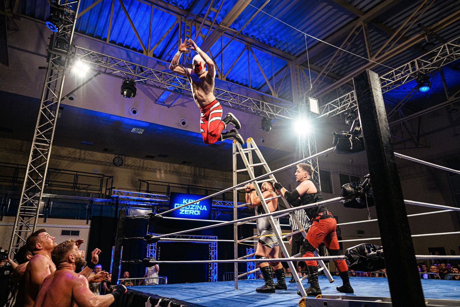 Wrestling match featuring aerial maneuvers under blue lighting in an indoor arena.