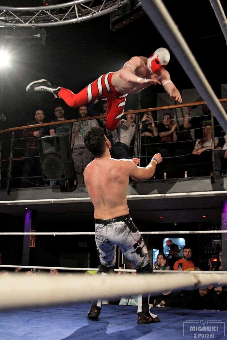 Wrestler executes a high-flying aerial move during a match in a dimly lit arena.