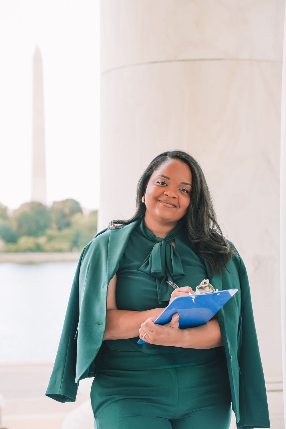 Professional headshot in green outfit with blue folder by white pillars and water view | Washington DC senior photos
